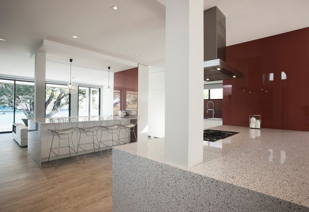Open-plan contemporary kitchen and living area with a large speckled stone island and matching foreground benchtop, glossy deep red splashback, white cabinetry and structural columns, timber-look flooring, pendant lights, and wire-frame bar stools. Floor-to-ceiling glazing opens the bright space to an outdoor setting.
