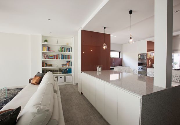A bright, open-plan contemporary kitchen and living area. A large white island with a speckled stone benchtop dominates the foreground, with pendant lights above. White full-height cabinetry and integrated appliances sit behind the island, while a white sectional sofa faces a built-in bookshelf filled with books. A deep red feature wall adds contrast to the otherwise white, light-grey, and neutral interior.
