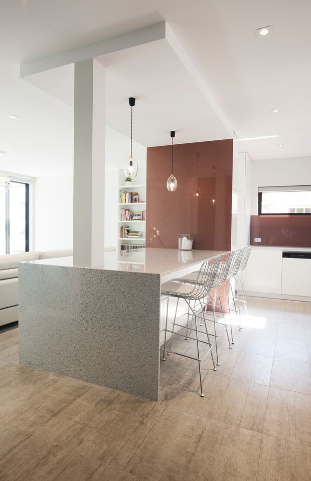 Bright open-plan contemporary kitchen and living area with a long island, pale speckled stone panels and benchtop, white cabinetry, wire-frame bar stools, pendant lights, and a glossy terracotta feature wall. A built-in bookshelf and partial sofa are visible beyond the island. The space has a clean, airy, minimalist composition with strong natural light.