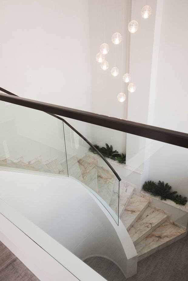A bright, high-ceilinged stairway viewed from an upper landing, featuring a sculptural curved white stair balustrade, clear glass panels with dark timber or metal handrails, pale veined stone stair treads, and a cluster of suspended spherical pendant lights. Built-in planter beds with greenery line the stairwell.