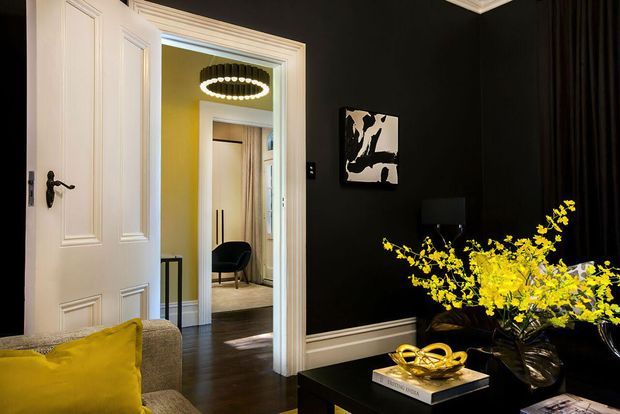 A dramatic, refined interior view of a black-painted living room with white traditional panelled doors, decorative cornices and skirting boards. A grey upholstered sofa with a mustard-yellow cushion sits beside a black coffee table styled with a sculptural gold ornament and bright yellow floral arrangement. Through the open doorway is a connected yellow-accented room with a pendant light, desk and chair.