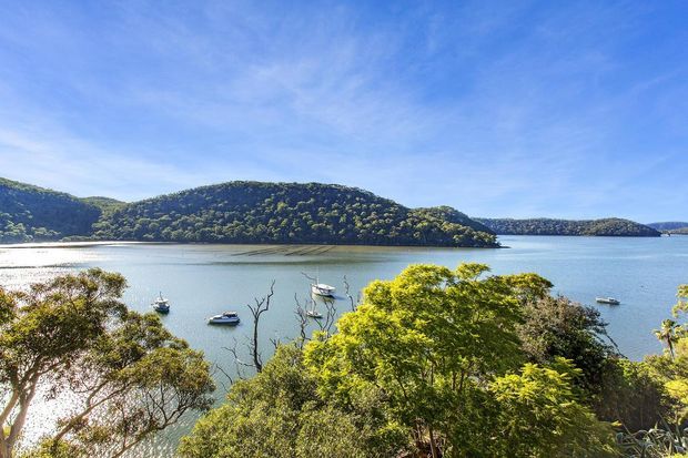 A bright scenic view across a calm coastal inlet, with several anchored boats, densely forested hills and islands, and leafy trees in the foreground beneath a blue sky.