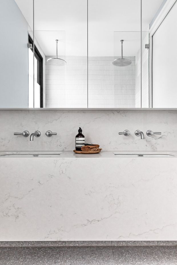 A bright, minimalist bathroom focused on a long double vanity with integrated white marble-look surfaces, wall-mounted chrome taps, and a large mirrored wall. A walk-in shower with fine white tiled walls and dual round rain showerheads is reflected in the mirror. The floor is finished in grey terrazzo tile.