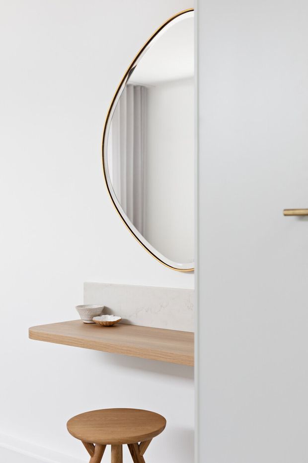 A bright, minimalist interior vignette featuring a round mirror with a thin brass-gold frame above a floating light timber shelf. Small ceramic vessels sit on the shelf, with a matching rounded timber stool below. A white door or partition with a slim brass handle partially frames the right side.
