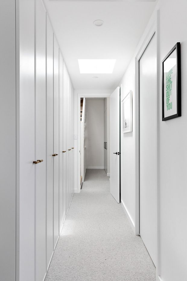 A bright, narrow residential hallway lined with tall white built-in storage cupboards and small timber handles on the left. White walls and ceiling, recessed lighting, a rectangular skylight or ceiling light, white doors with dark hardware, framed artwork, and a light grey carpet create a clean, minimalist corridor.
