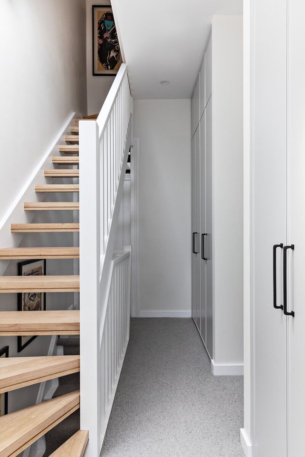 A bright, narrow upstairs hallway beside an open-riser staircase. The staircase has pale timber treads, white stringers and vertical white balustrades, while the hallway includes full-height white built-in cabinetry with black handles and a light grey carpet. Framed artwork is visible near the stair landing. The overall design is clean, contemporary and minimal.