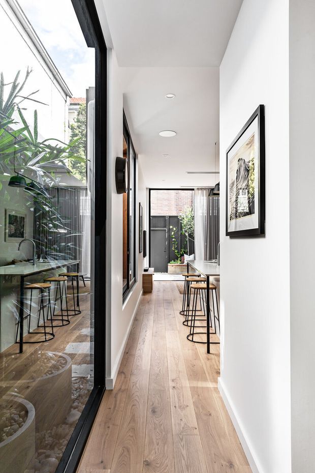 A bright, narrow contemporary interior corridor with white walls, pale timber flooring, black-framed full-height glazing, framed artwork, and a compact dining or kitchen area visible at the end. The glazing reflects a planted courtyard with an outdoor table and stools, while the corridor includes simple modern lighting and bar stools along a counter.