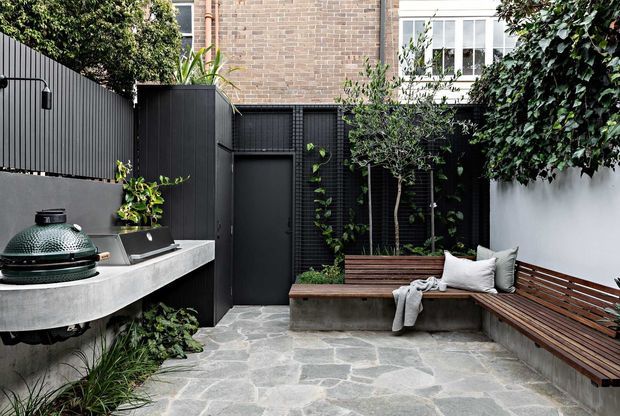 A compact, enclosed urban courtyard with dark vertical cladding, a built-in outdoor kitchen and barbecue, irregular pale stone paving, raised timber planter seating, and a long built-in timber bench with cushions. Dense greenery softens the black and white walls, while exposed brick and windows of the surrounding building are visible above.