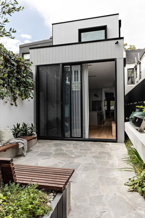 Contemporary two-storey rear extension viewed from a landscaped courtyard. The facade combines pale vertically ribbed cladding with dark window and door frames, large sliding glazing, and a flat roofline. A light irregular stone-paved patio leads into an open-plan interior, with timber bench seating, planted garden beds, and an outdoor barbecue area.