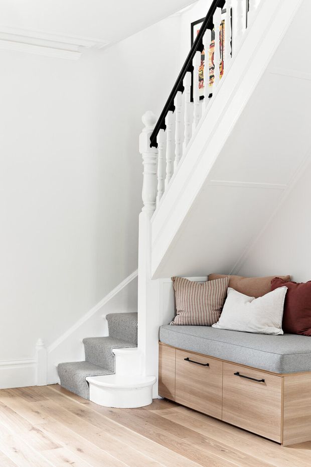 Bright white stairway with traditional turned balusters and a black handrail, leading beside a compact built-in window seat or storage bench beneath the stairs. The space features white paneled walls and trim, pale timber flooring, light wood cabinetry with black pulls, and neutral upholstered cushions.