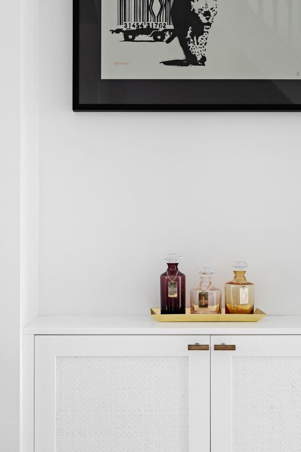 A close-up of a refined white built-in cabinet or console with textured inset door panels and slim brass-toned handles. A small brass tray holding decorative glass perfume bottles sits on the countertop, while a black-framed monochrome artwork hangs above. The composition is minimal and monochromatic with warm metallic accents.