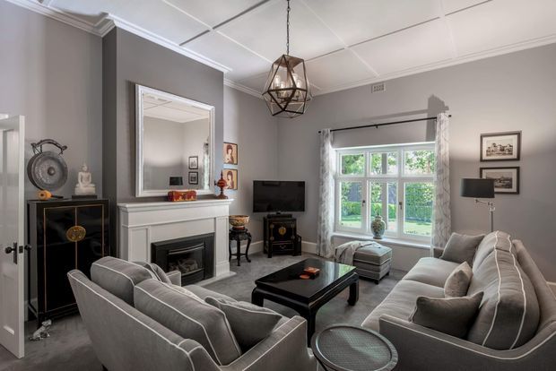 A refined living room with grey upholstered sofas arranged around a black coffee table, a white fireplace mantel with mirror, a television, framed artwork, and a large multi-pane window with curtains. The room features decorative ceiling mouldings, a coffered ceiling, and a geometric pendant light, creating a polished traditional-contemporary interior.