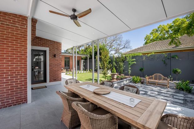 A covered outdoor dining terrace beside a red-brick home, furnished with a large timber dining table and woven wicker chairs. The space has a white-lined ceiling with a timber-bladed ceiling fan, tiled flooring, slender white posts, and views onto a landscaped courtyard with dark fencing, gravel, planting, and a bench.