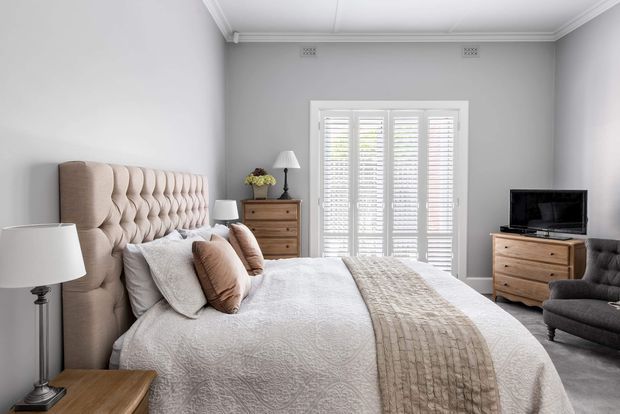 A bright, refined bedroom with a large upholstered button-tufted headboard, layered neutral bedding, bedside tables and lamps, timber drawer chests, plantation shutters, and a grey upholstered armchair. The room has pale grey walls, white decorative ceiling mouldings, and soft natural light.