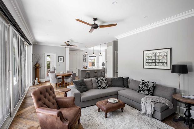 Bright open-plan living room with a large grey sectional sofa, tufted leather armchair, ottoman, patterned cushions, light rug, and framed wall artwork. Full-height glazed doors line one side, while the rear connects to a dining area and kitchen with island seating. White walls, decorative ceiling cornices, timber flooring, pendant lights, and ceiling fans create a refined, comfortable interior.