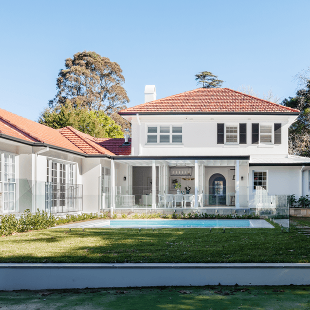 A bright exterior view of a substantial white two-storey residence with a terracotta tiled hipped roof, classical shuttered windows, covered verandah, and glazed side wings. A rectangular swimming pool and manicured lawn occupy the foreground, with mature trees behind the house.