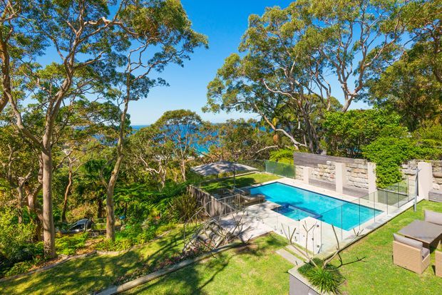 A bright aerial view of a landscaped residential property set among mature trees, featuring a turquoise swimming pool with glass balustrades, paved pool terrace, outdoor seating, and a rendered masonry privacy wall. Blue water is visible beyond the treetops, suggesting a coastal or waterfront setting.