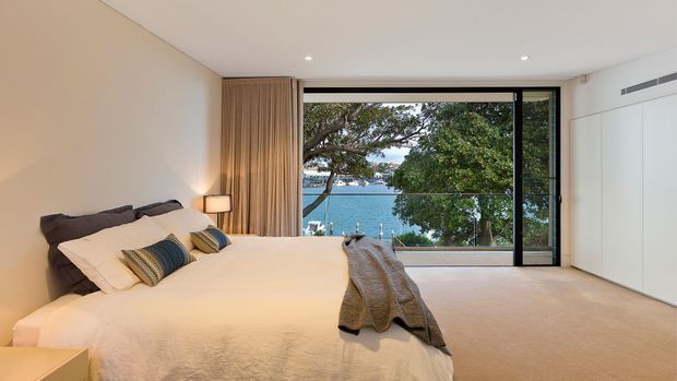 A bright, contemporary bedroom with a large bed dressed in white bedding, textured neutral cushions, and a grey throw. Full-height timber curtains frame expansive black-framed glazing overlooking water and trees. The room has soft beige carpeting, white walls, integrated white cabinetry, recessed ceiling lighting, and a wall-mounted air-conditioning vent.