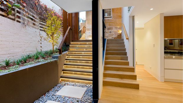 A contemporary split-level residential interior showing an illuminated timber staircase beside a landscaped internal courtyard. The view includes a white-painted brick garden wall, pebble groundcover, planted greenery, timber screens and stairs, exposed stonework, and an adjoining light timber-floored kitchen area with warm wood cabinetry.