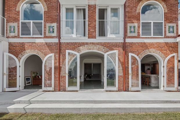Exterior view of a multi-storey red-brick building with symmetrical arched openings, white-framed windows and doors, louvred shutters, and broad concrete steps leading to several open ground-floor entries. Interior spaces are visible through the doors, including a small bar or kitchenette area on the right.