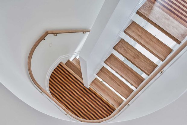 Top-down view of a curved stairway with open timber treads and a matching curved timber handrail. The staircase has a white painted structure and walls, with strong geometric lines and visible light passing between the treads.
