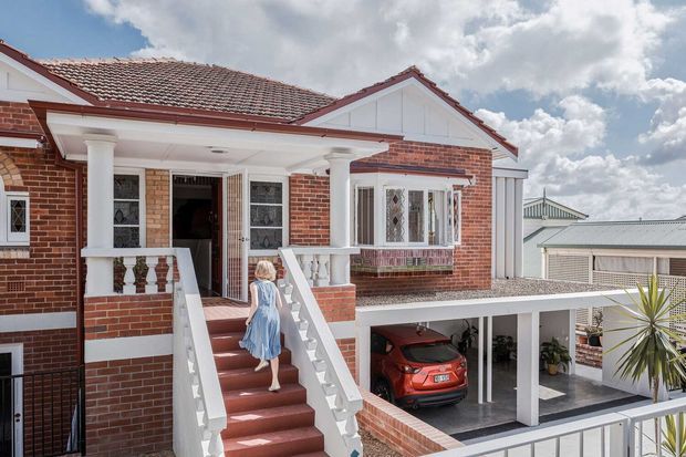 Exterior view of a traditional brick house with a red tiled gable roof, white-painted trim, columns, balustrades, and a raised front entry accessed by a staircase. A covered carport and red car are visible below, with a neighboring roof in the background.