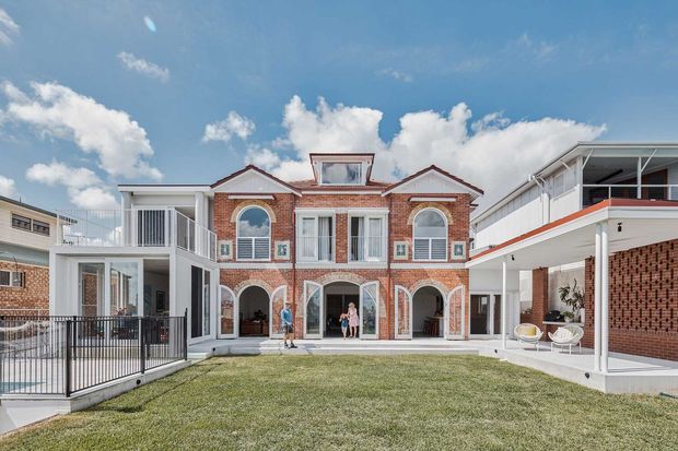 Wide exterior view of a substantial red-brick residence arranged around a landscaped courtyard. The central two-storey facade features arched openings, white classical trims, decorative gable details and tall multi-pane windows, flanked by contemporary white-clad wings with balconies and covered outdoor areas. The image is captured in bright daylight with a blue sky and lawn in the foreground.