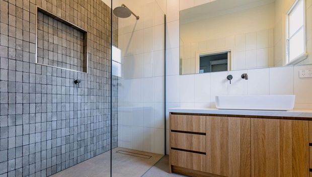 Contemporary bathroom featuring a frameless glass walk-in shower, grey square tiled shower wall with recessed niche, white tiled walls, and a timber-look floating vanity with a white vessel basin and wall-mounted black fixtures. Natural light enters through a side window.