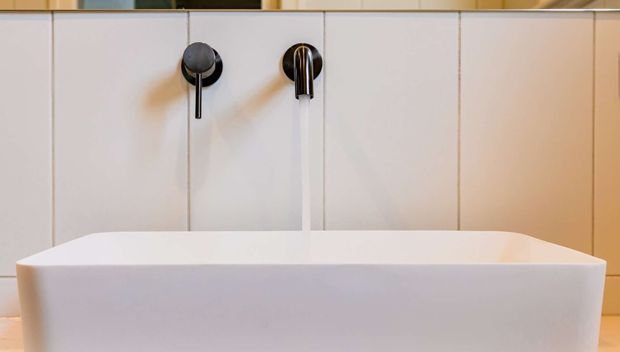 Close-up of a contemporary freestanding white bathtub with a wall-mounted black and chrome faucet set. Large pale wall panels form a clean backdrop, with warm wood-toned cabinetry or trim visible at the edges.