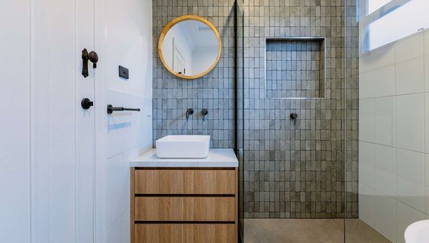 Compact contemporary bathroom with a timber floating vanity, white vessel basin, round brass-framed mirror, dark metal tapware, and a walk-in shower lined with small textured grey tiles. White wall panelling and pale floor tiles create a clean, light finish.