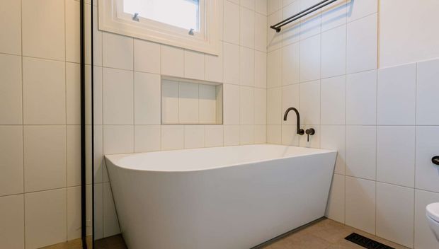 A clean, contemporary bathroom featuring a freestanding white bathtub, white tiled walls, a recessed wall niche, a small high window, dark tapware, a wall-mounted towel rail, and warm beige floor tiles. A partial toilet is visible at the right edge.