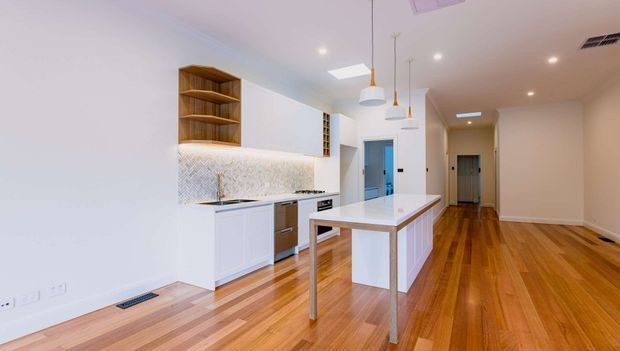 Bright open-plan kitchen and living/dining space with white cabinetry, a long central island, timber flooring, pendant lighting, and integrated open shelving. The kitchen features a patterned tiled splashback and warm timber accents, with a hallway extending toward the rear.
