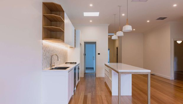 Bright contemporary kitchen with white cabinetry, a large white island, warm timber flooring, open timber shelving, a light tiled backsplash, and pendant lighting. The kitchen opens toward a hallway and adjacent living area.