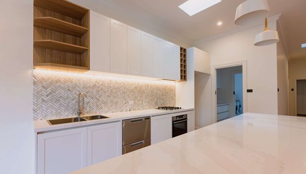 Bright contemporary kitchen with white shaker-style cabinetry, warm timber open shelving, a white engineered-stone island and benchtops, and a light herringbone tile splashback. Integrated appliances, a stainless-steel sink and tap, under-cabinet lighting, pendant lights, and a skylight create a clean, refined fit-out.