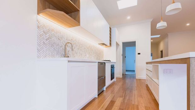 Bright contemporary galley kitchen with white panelled cabinetry, a pale herringbone tile splashback, warm timber open shelving, timber flooring, integrated appliances and a peninsula bench. Pendant lights and recessed ceiling lighting illuminate the elongated space, with a hallway visible beyond.