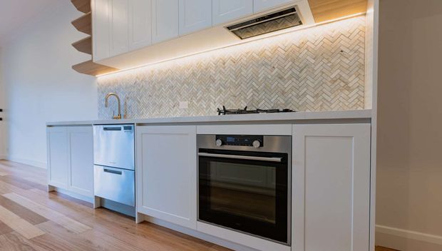 Compact contemporary kitchen with pale blue-grey shaker-style cabinetry, integrated stainless-steel dishwasher, built-in black oven, gas cooktop, and a light herringbone tile splashback. Warm under-cabinet lighting and timber flooring add contrast, while a sculptural stepped open shelf is visible at the left.