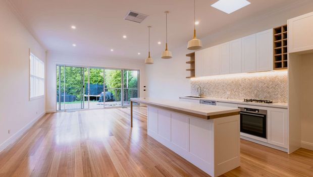 Bright open-plan kitchen and living area with a large white island, integrated cabinetry, patterned tile splashback, timber flooring, pendant lights, and full-height glazed doors opening to a leafy outdoor area.