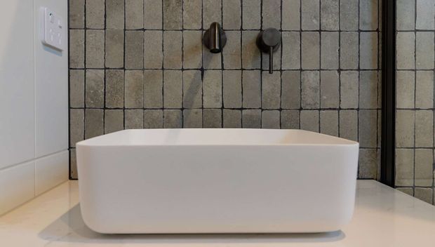 A modern bathroom featuring a freestanding white bathtub positioned against a textured dark-grouted tiled wall, with wall-mounted dark metal tapware and a pale tiled floor.
