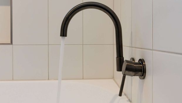 Close-up of a dark metal wall-mounted gooseneck bath tap with water running into a white bathtub. The surrounding walls are finished with large-format light neutral square tiles and matching grout, creating a clean, understated bathroom setting.
