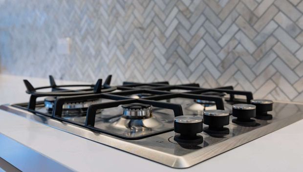 Close-up view of a stainless-steel gas cooktop with multiple black control knobs and cast-iron pan supports, set into a light countertop. A pale grey herringbone tile splashback forms the background.