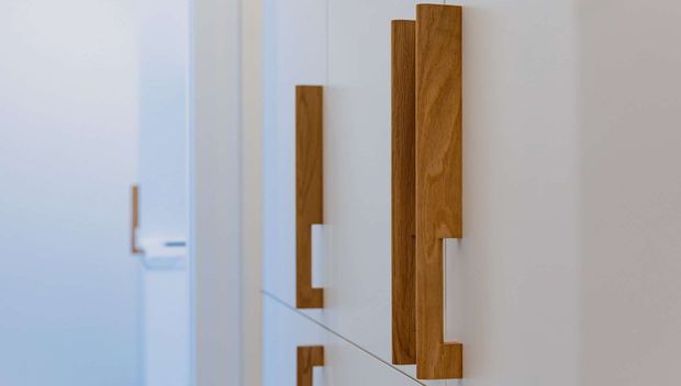Close-up of a minimalist interior wall featuring a series of tall, sculptural timber door or cabinet handles mounted on a smooth white surface. The warm wood contrasts with the cool white background, while a faint reflection or glass edge adds depth.