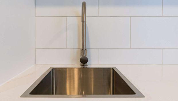 Close-up of a modern kitchen sink with a deep stainless-steel undermount basin and tall brushed-metal gooseneck tap, set against a white subway-tile backsplash and pale benchtop.