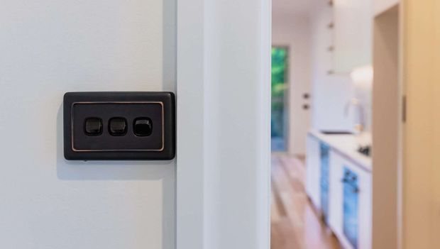 Close-up of a dark three-button electrical switch plate mounted on a white wall beside a doorway. A bright, predominantly white kitchen with light timber flooring and cabinetry is softly visible through the opening in the background.