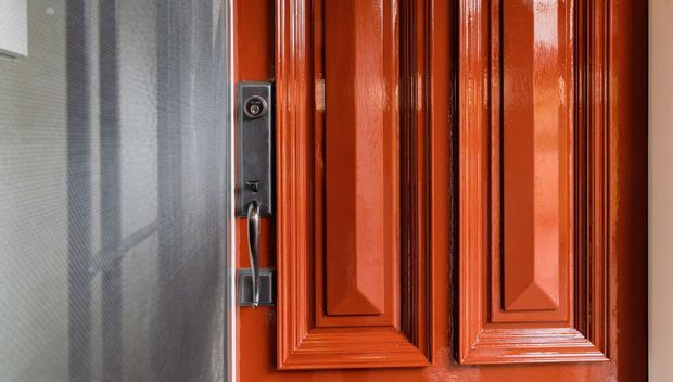 Close-up of a glossy reddish-orange traditional paneled exterior door with raised moulded detailing and dark metal lock hardware, viewed beside a translucent mesh security screen door.