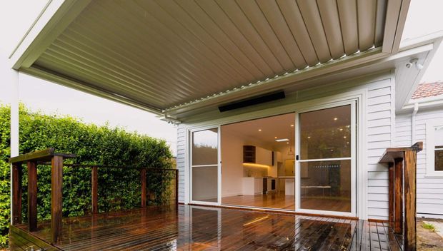 A contemporary covered outdoor deck adjoining a white weatherboard house. The deck has glossy timber decking, a large adjustable louvred pergola roof, timber privacy screens and balustrades, and wide sliding glazed doors opening into a warmly lit open-plan interior. A compact kitchen or living area is visible inside.
