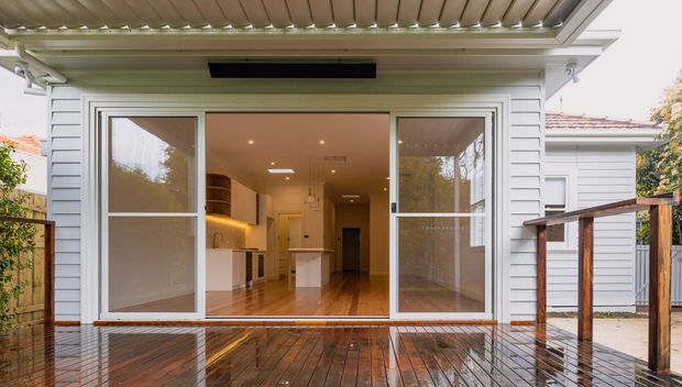 Rear exterior view of a renovated white weatherboard home opening onto a wet timber deck. Large sliding glazed doors reveal a warm, modern open-plan kitchen and living area, while a covered pergola and timber balustrade frame the outdoor space.
