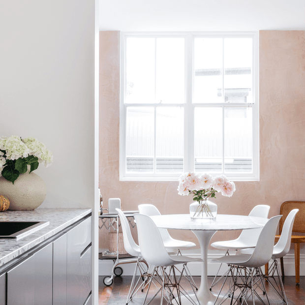 Bright contemporary dining area adjoining a modern kitchen. The room features a white round pedestal dining table, several white molded chairs, a vase of pale flowers, large white-framed windows, pale pink textured wall panels, and polished light-toned cabinetry with a stone-look benchtop in the kitchen at left.