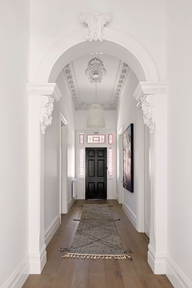 A narrow, elegant residential hallway viewed through a large ornate arched opening. The space has white walls and elaborate classical plaster detailing, including fluted architraves, carved corbels, decorative ceiling mouldings and a central ceiling medallion. A black panelled entry door is framed by decorative leadlight glazing at the far end. Wide-plank timber flooring, a long neutral patterned runner, a woven pendant light and framed artwork complete the refined, heritage-inspired interior.