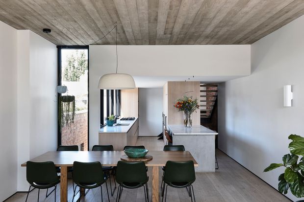 A bright open-plan dining and kitchen interior. A timber dining table with dark green dining chairs occupies the foreground, while a compact kitchen with pale wood cabinetry, a light stone island benchtop, integrated appliances and a small pantry or shelving area sits beyond. The room features white walls, a full-height window, grey-toned timber flooring and a striking timber-lined ceiling. A large neutral pendant light hangs above the dining table, with indoor planting adding greenery.