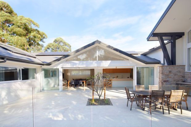 A bright contemporary courtyard centered on a glazed, gable-roofed pavilion with an open-plan kitchen and dining area. Pale tiled or concrete-look paving extends across the courtyard, with timber outdoor dining furniture, a small planted tree, and adjoining modern volumes with stone-clad walls and dark roof framing.